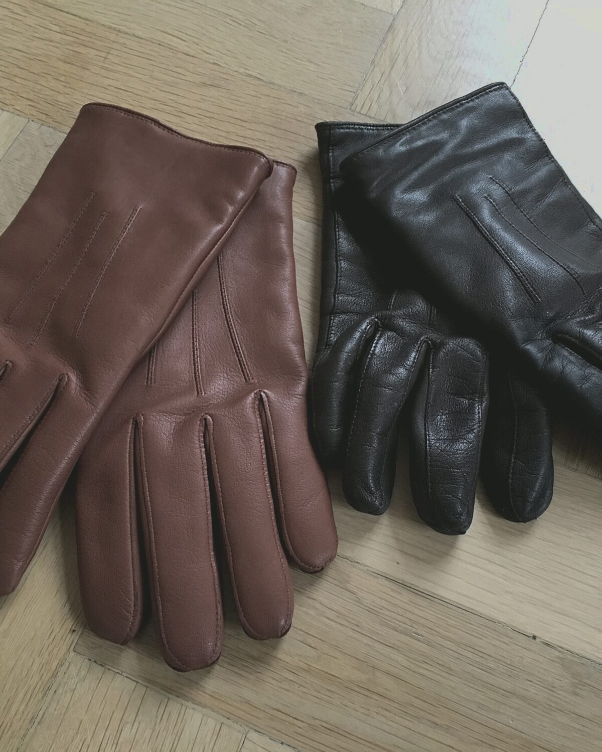 Brown peccary driving gloves beside a black pair, laid on a pale surface